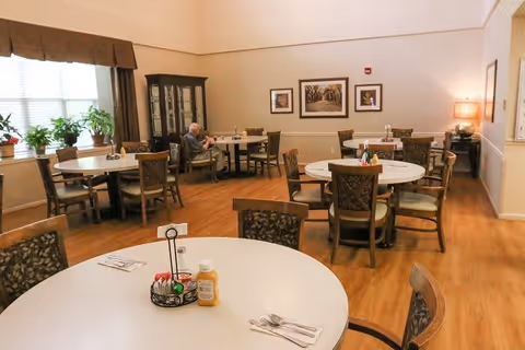 A dining room in a senior living facility with several round tables and chairs arranged neatly. One elderly person is seated at a table near the window. The room has wooden flooring, framed pictures on the wall, a cabinet with glass doors, and a lamp on a side table. There are plants on the windowsill and condiments on the tables.