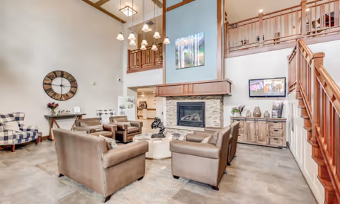 A spacious senior living facility common area with high ceilings, a stone fireplace, and a seating arrangement consisting of four brown leather armchairs around a round coffee table. There are two plaid armchairs near a wall with a large decorative clock. A wooden staircase with railings leads to an upper level balcony. The walls are decorated with artwork and a flat-screen TV is mounted above a wooden cabinet.