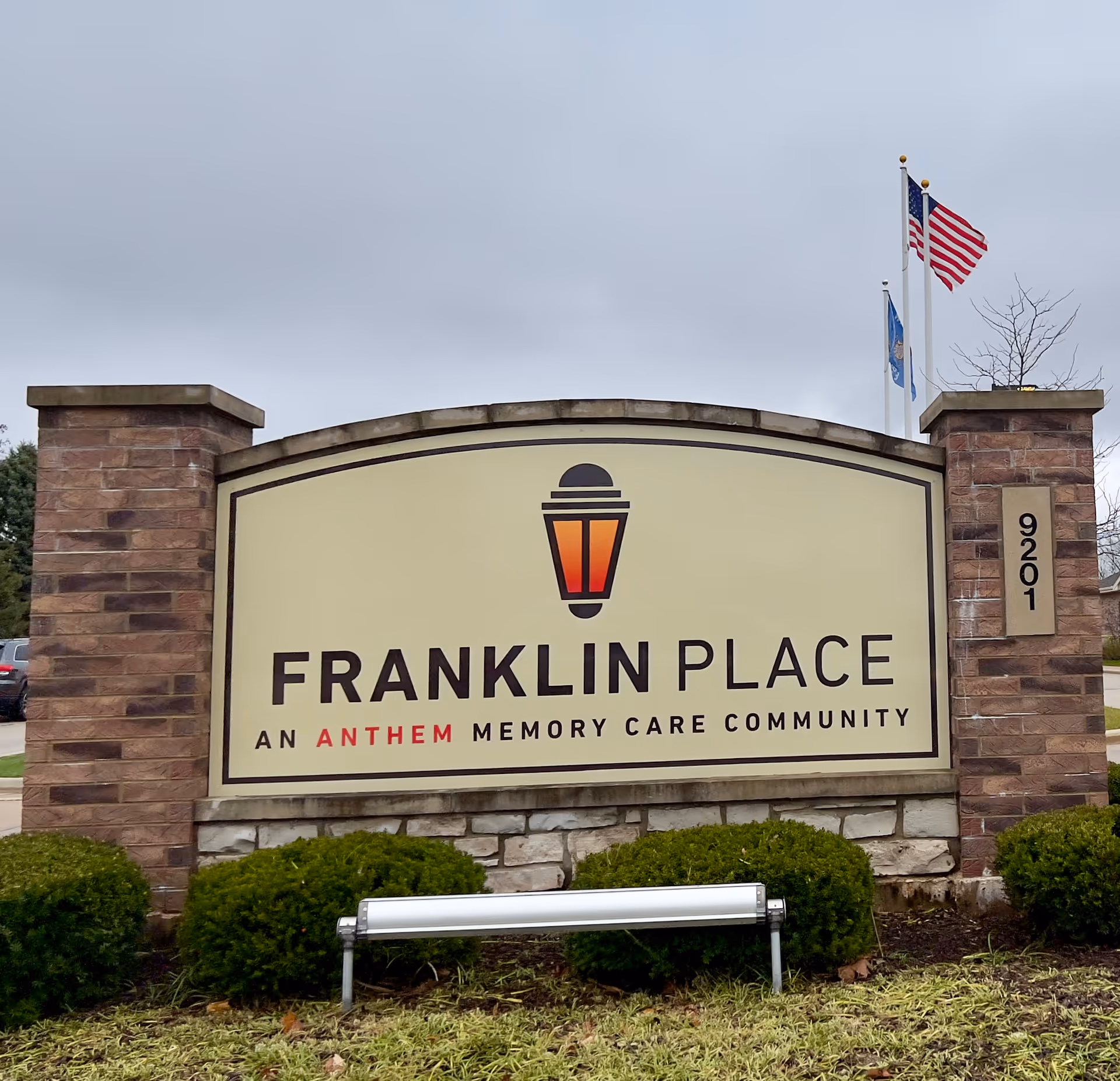Outdoor sign for Franklin Place, an Anthem Memory Care Community, mounted on a stone and brick structure with two flagpoles flying the American flag and another flag in the background under a cloudy sky.