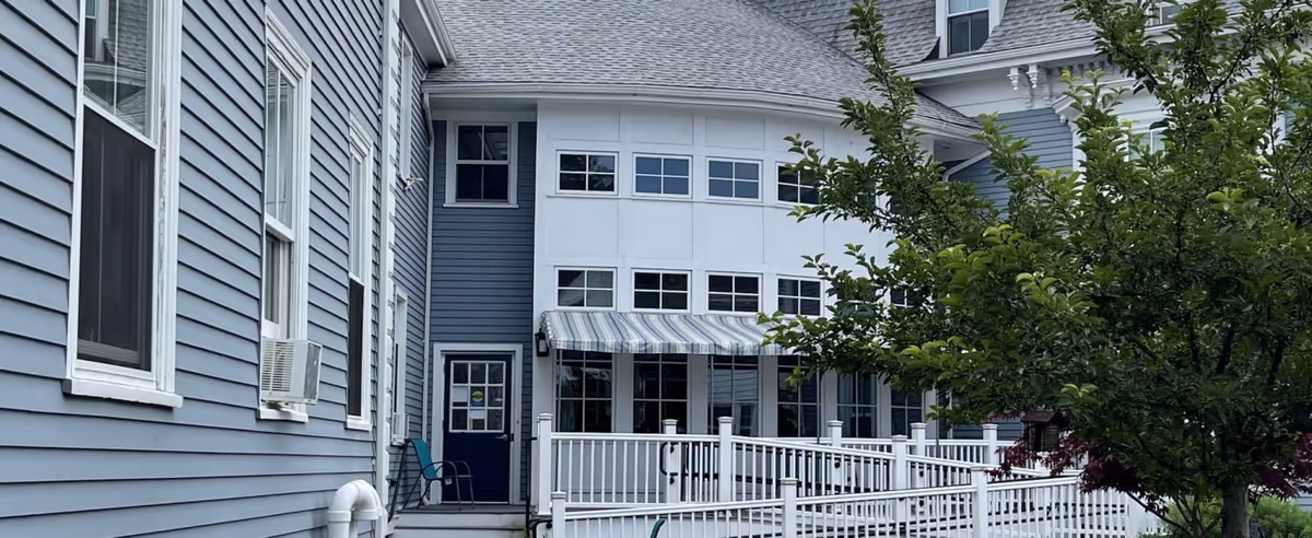 Exterior view of a blue senior living building with a white railing deck and striped awning over the entrance.