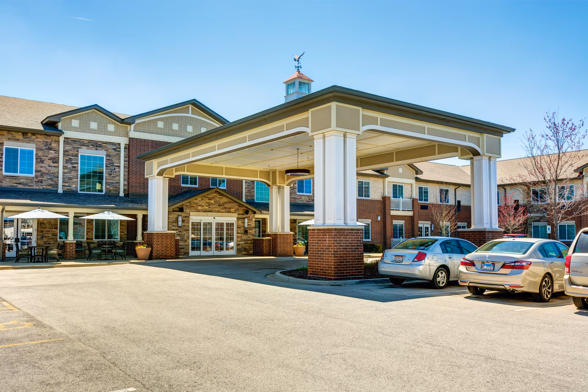 Exterior view of Burr Ridge Senior Living facility showing the main entrance with a covered drop-off area supported by large white columns with brick bases. Several parked cars are visible in front of the building, which features a mix of brick and stone facade with multiple windows. Outdoor seating with umbrellas is seen to the left side of the entrance under a clear blue sky.