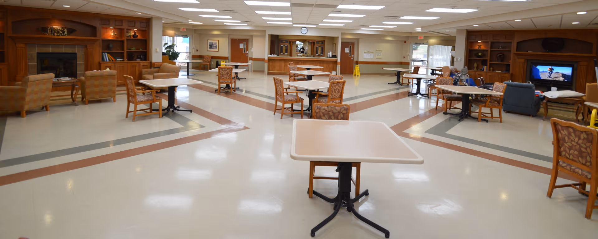 A spacious common area in a senior living facility with multiple tables and chairs arranged throughout the room. There are comfortable armchairs near a wooden fireplace and built-in shelves. A television is on in the corner, and large windows allow natural light to enter. The room has a clean, polished floor with decorative tile patterns and a bright ceiling with recessed lighting.