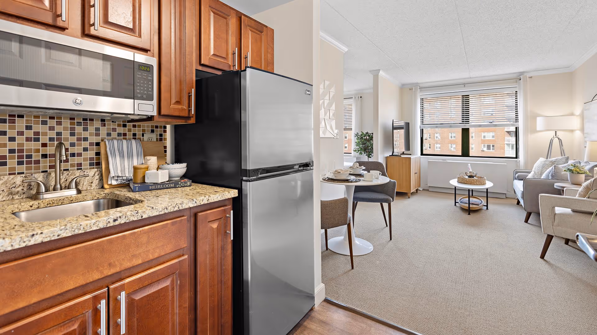 A bright and clean interior space featuring a small kitchen area with wooden cabinets, a granite countertop, a stainless steel sink, microwave, and refrigerator. Adjacent to the kitchen is a cozy living room with a round dining table set for two, a sofa, armchairs, a coffee table, a TV on a stand, and large windows letting in natural light.