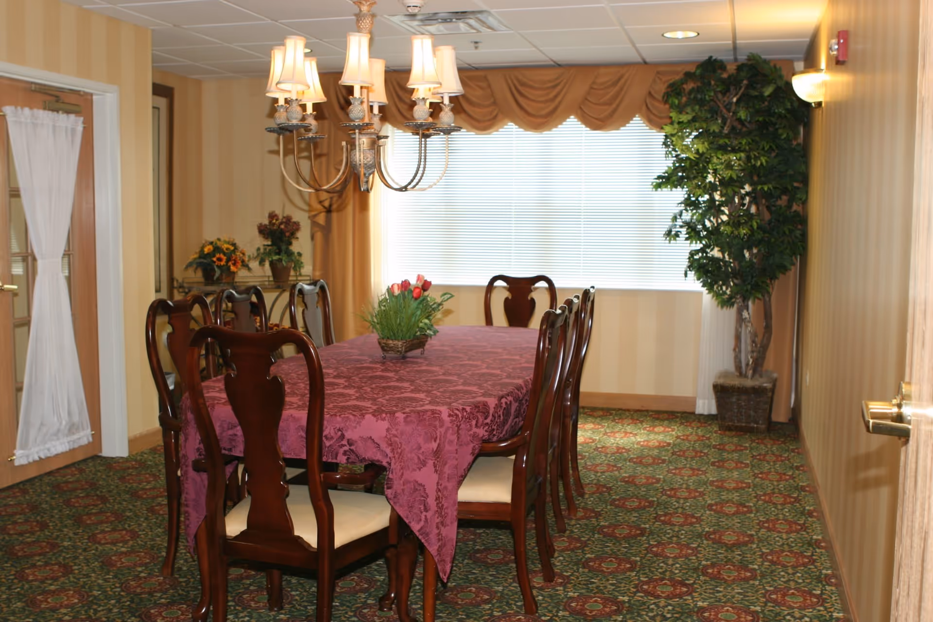Formal dining room with a long table covered by a purple tablecloth, wooden chairs, chandelier, and a window with blinds.