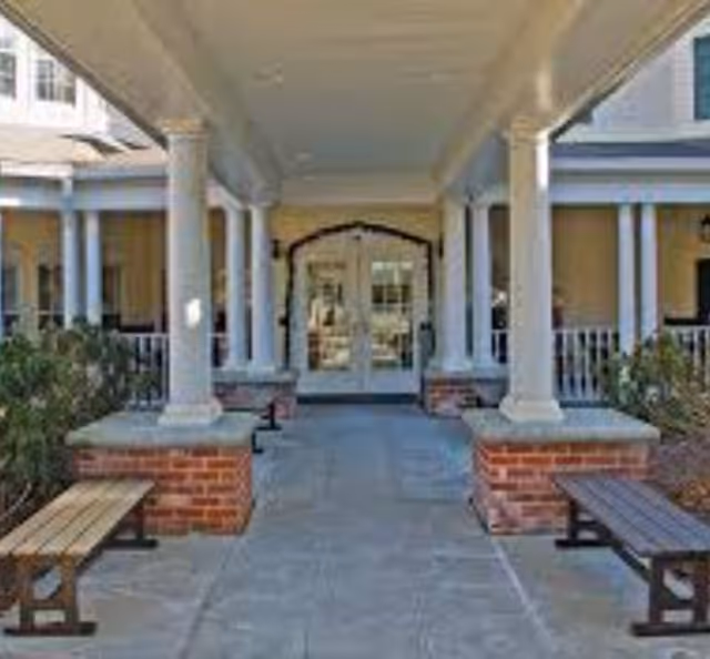 Covered entrance area of a building with white columns, brick bases, two wooden benches on either side, and glass double doors at the far end.