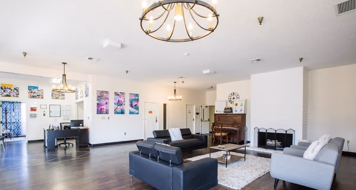 A spacious and well-lit living room area in a senior living facility with modern black and gray sofas arranged around a coffee table on a white rug. There is a white brick fireplace on the right side and a wooden piano with a chair next to it. The walls are decorated with colorful artwork and a large clock. The room has dark wooden flooring and two large chandeliers hanging from the ceiling. In the background, there is a desk with a computer and some office supplies near an entrance with a patterned curtain.