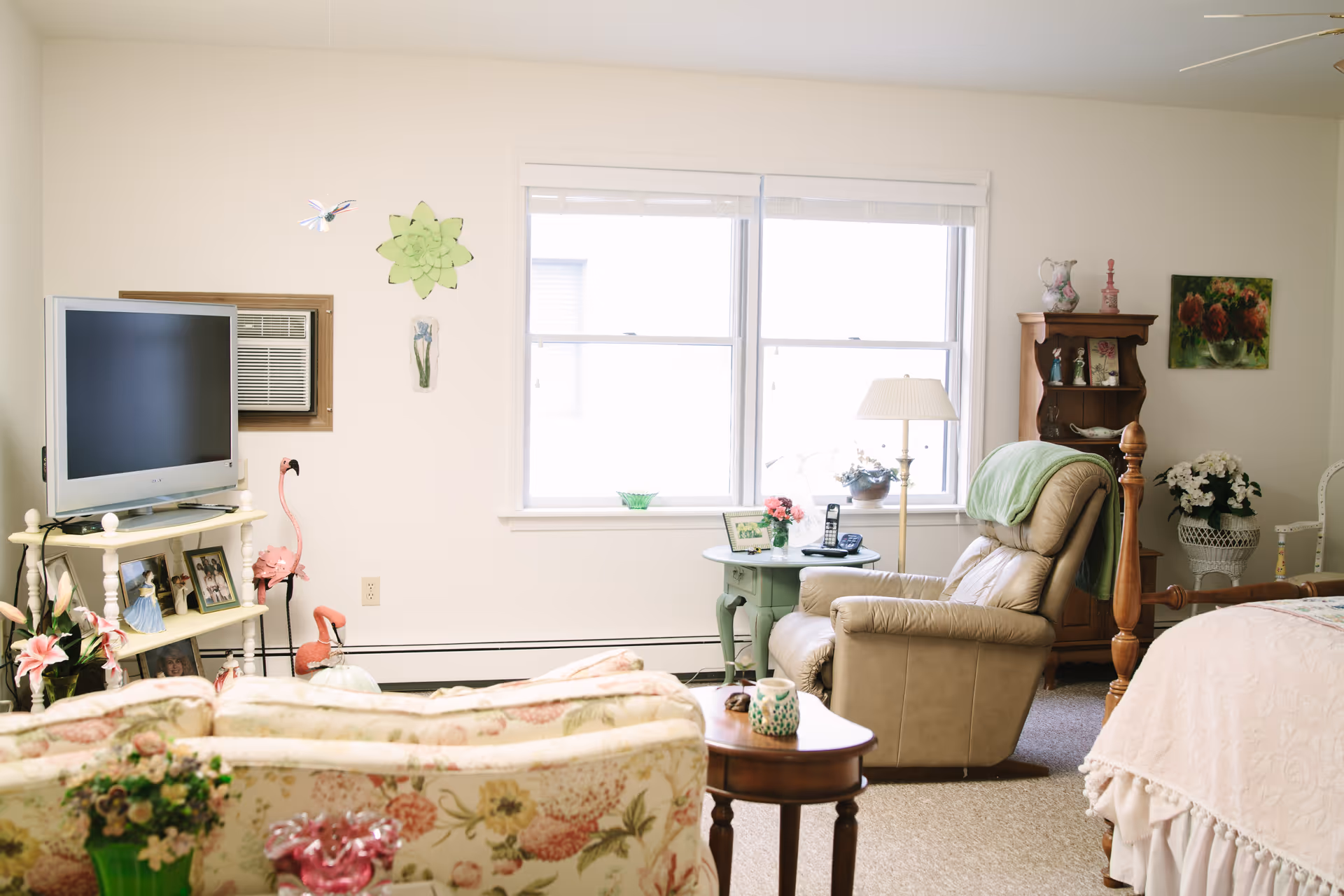 A cozy senior living room and bedroom area with a floral patterned sofa, a beige recliner chair, a wooden side table, and a bed with a light pink quilt. The room has a window with blinds, a TV on a white stand, an air conditioning unit, and various decorative items including flowers, framed photos, and wall art.