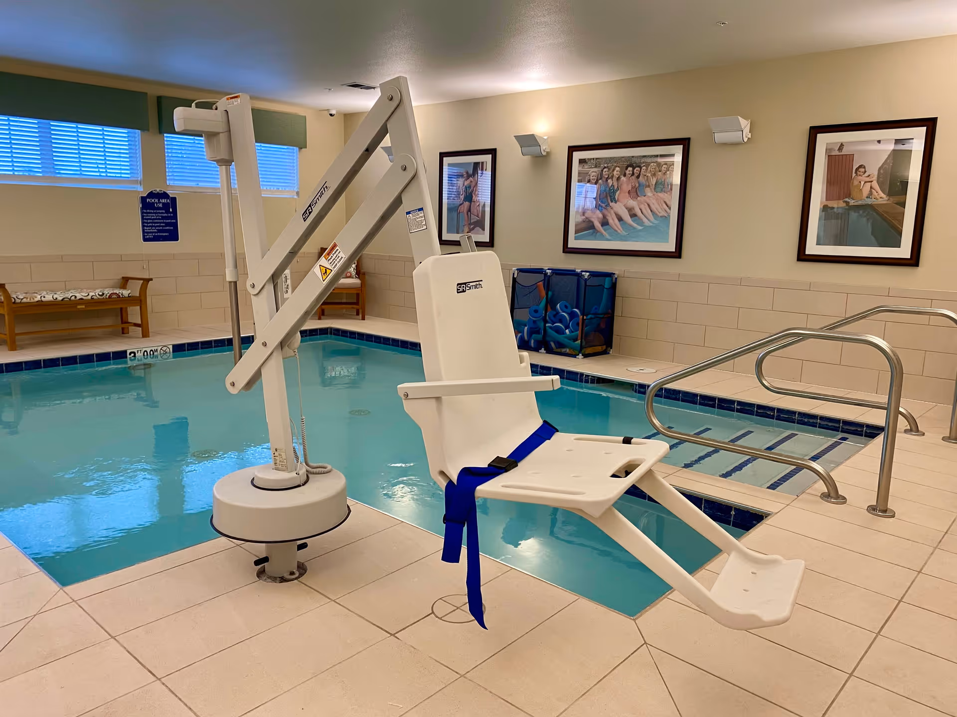 Indoor swimming pool area with a white pool lift chair for accessibility, beige tiled floor, metal handrails leading into the pool, and framed pictures on the wall. There is a bench and a basket with pool noodles in the background.