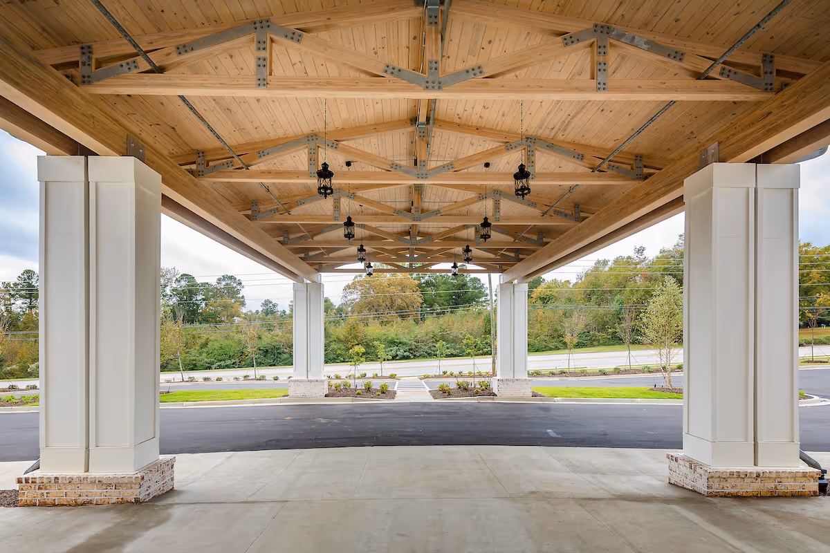 Covered entrance area with a wooden ceiling supported by large white columns with brick bases, overlooking a driveway and landscaped greenery with trees in the background.