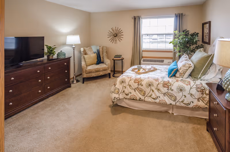 A cozy bedroom with a patterned bedspread on a bed, several pillows, a wooden nightstand with a lamp, a large window with curtains, a cushioned armchair with a pillow and throw, a small round side table, a wooden dresser with a flat-screen TV and plants, and beige carpeted floor.