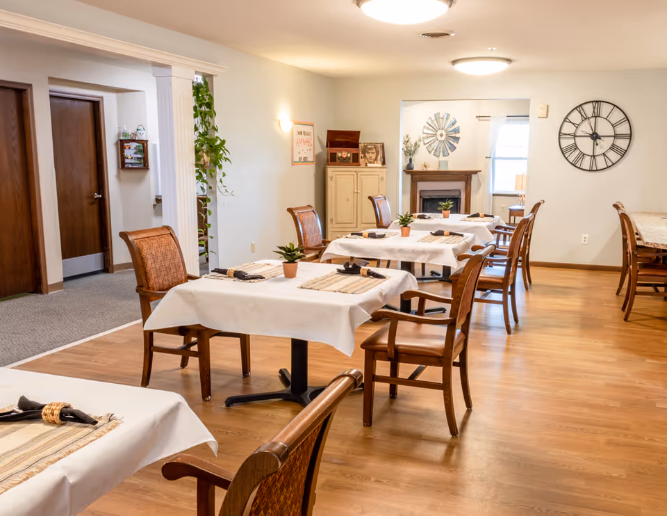 A dining room in a senior living facility with several tables covered in white tablecloths and set with placemats, napkins, and small potted plants. Wooden chairs with cushioned seats surround the tables. The room has wooden flooring, a large wall clock, a fireplace, and a cabinet with decorative items. There is a window with curtains letting in natural light.