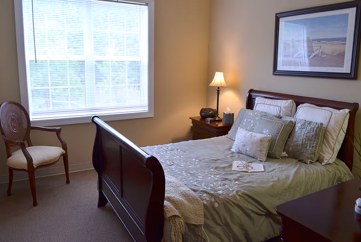 A cozy bedroom with a wooden bed frame, neatly made bed with multiple pillows, a bedside table with a lamp, a chair near a large window with blinds, and a framed picture of a beach scene on the wall.