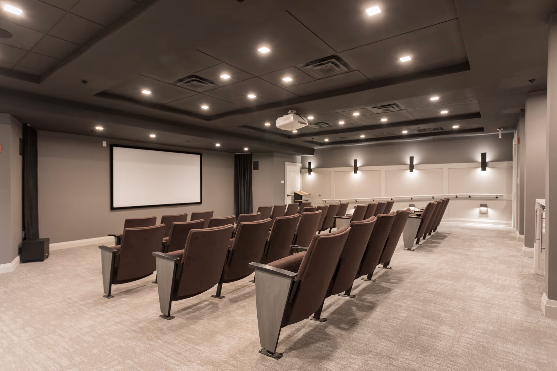 A modern theater room with rows of brown cushioned seats facing a large blank projection screen on the wall. The room has a gray ceiling with recessed lighting and a projector mounted in the center. The walls are painted gray with white trim and wall sconces providing additional lighting. The floor is carpeted in a light beige color.