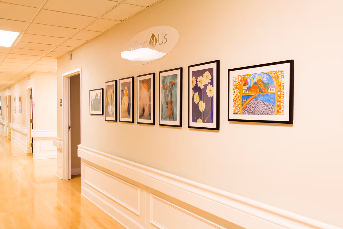 A well-lit hallway in a senior living facility with light-colored walls and wooden flooring. Several framed artworks are displayed on the right wall, and there are multiple doorways along the left side of the hallway. A sign above the artwork reads 'Reflections'.