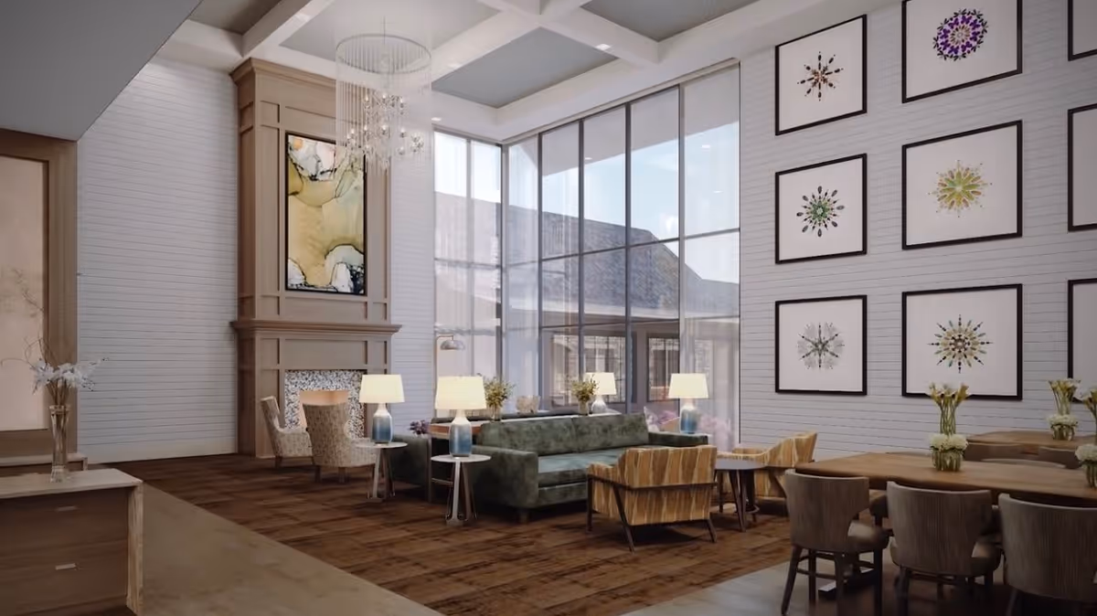 A spacious and well-lit living room area with large floor-to-ceiling windows, a green sofa, several armchairs, side tables with lamps, a fireplace with artwork above it, and a wall decorated with framed abstract art pieces. There is also a wooden dining table with chairs and vases with flowers on the tables.