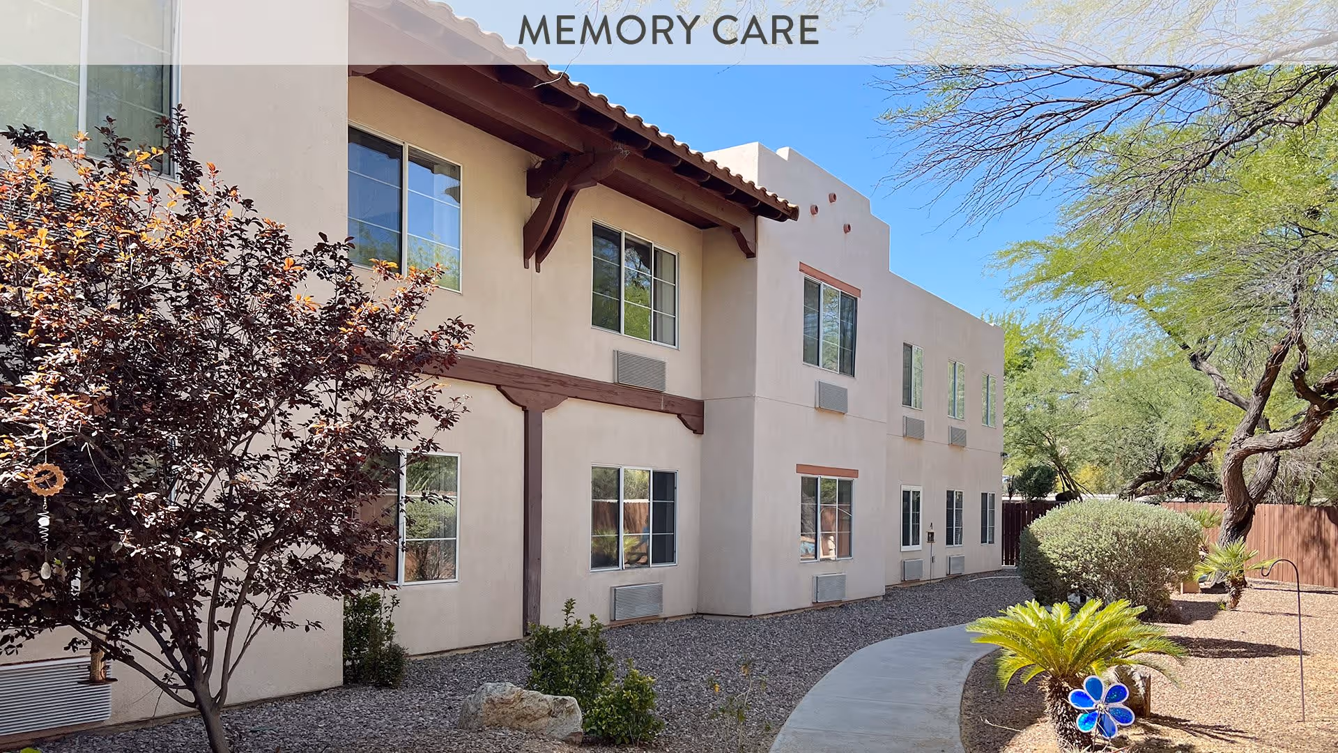Exterior view of a two-story beige building with multiple windows, surrounded by a landscaped garden with trees, bushes, and a curved concrete pathway under a clear blue sky. The words 'MEMORY CARE' are visible at the top of the image.