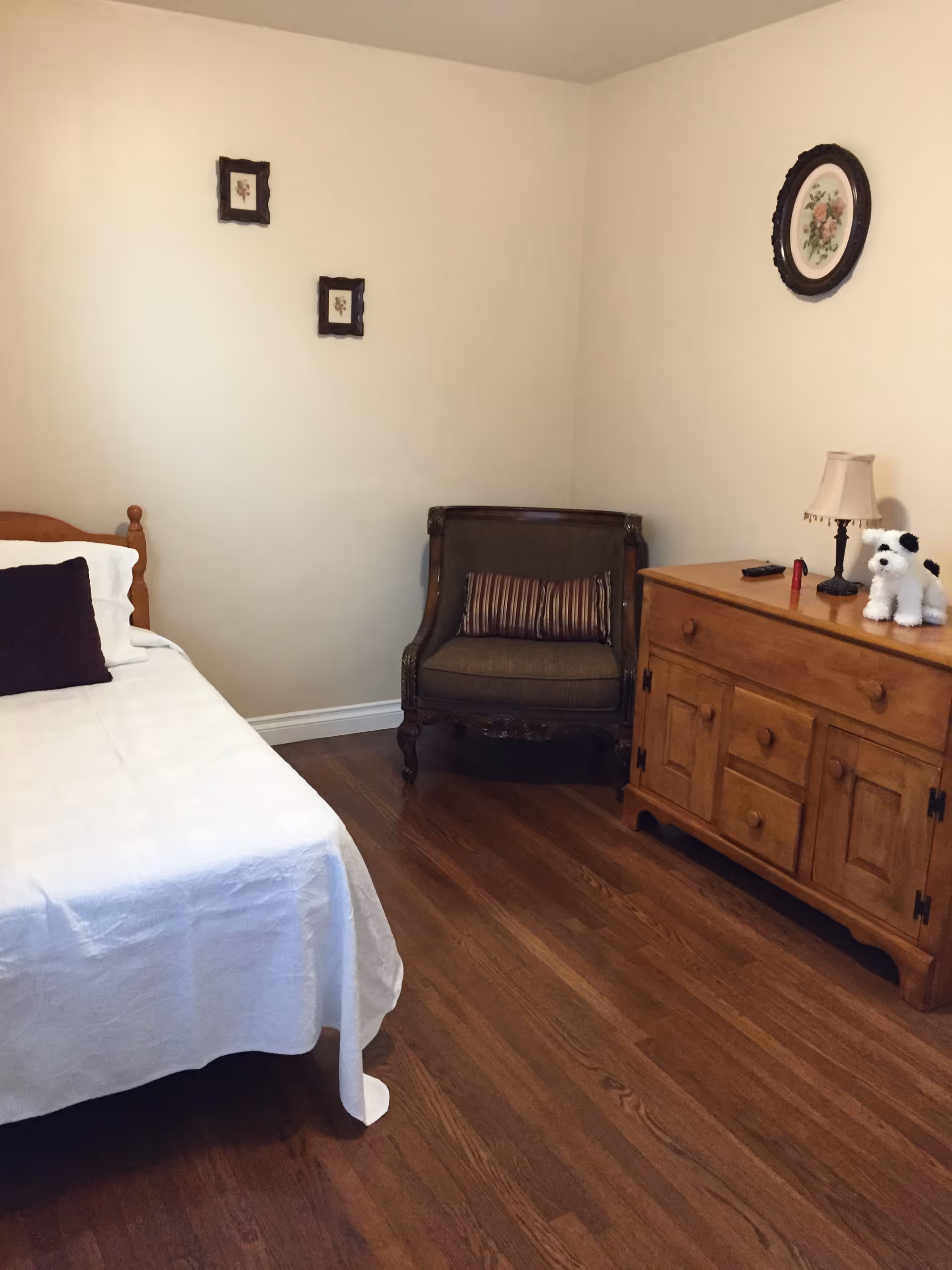 Small bedroom with a single bed, wooden dresser, armchair and framed wall decorations.