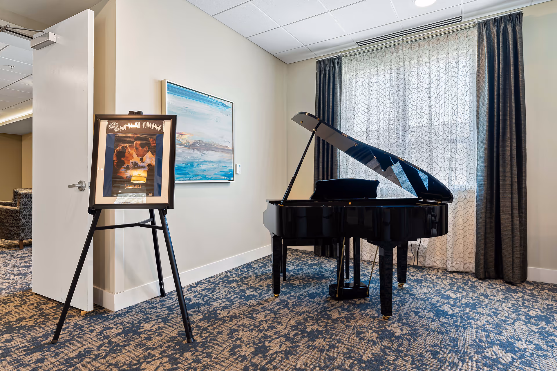 A black grand piano sits in a carpeted common room beside a framed poster on an easel and a window with patterned curtains.