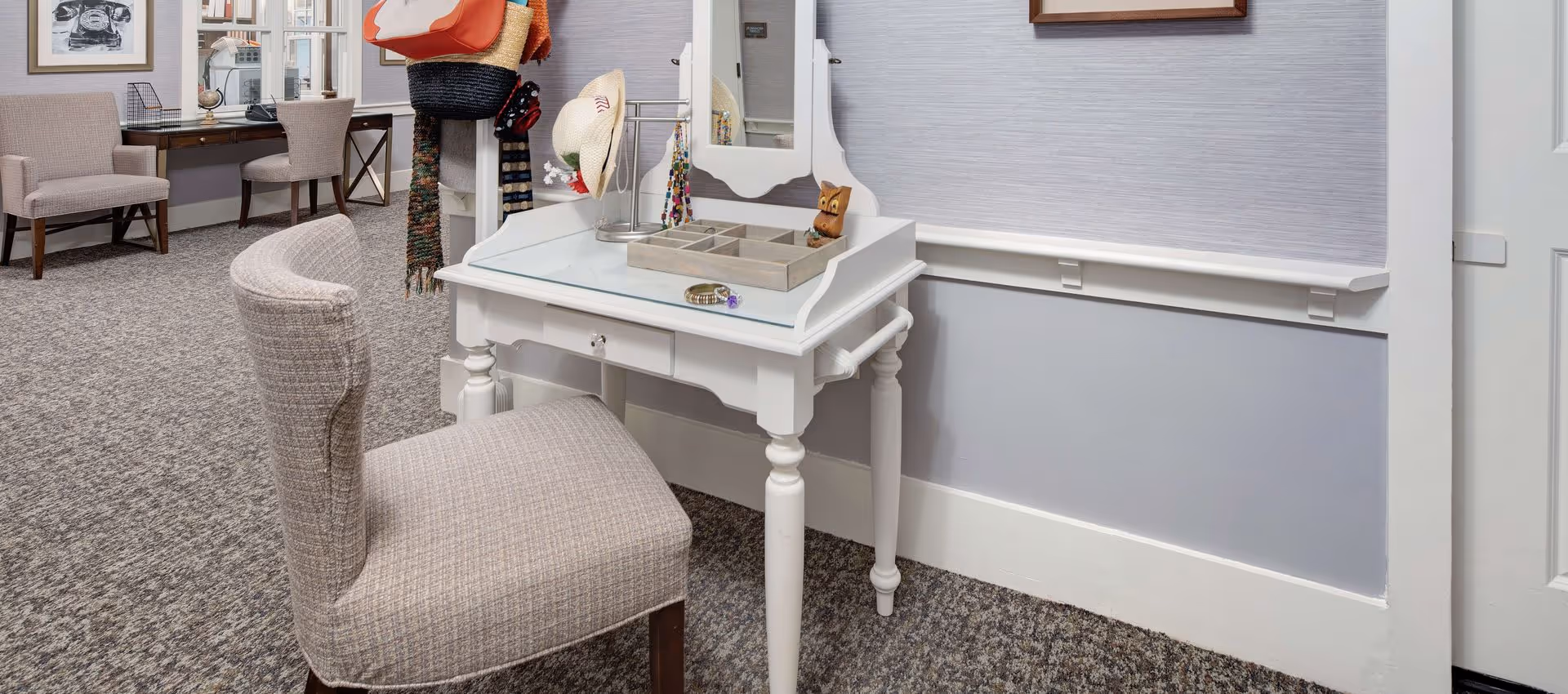 A cozy interior space featuring a white vanity table with a mirror and a cushioned chair. The vanity has a glass top with a jewelry organizer and a bracelet on it. There are hats and scarves hanging on a stand nearby. In the background, there is another chair and a desk with a globe and a vintage telephone on it. The walls are light gray with white trim, and the floor is carpeted in a textured pattern.