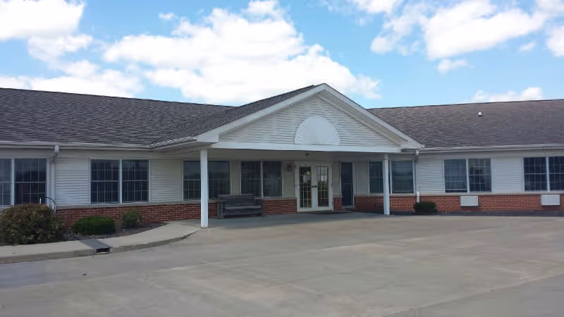 Front exterior view of a single-story building with white siding and red brick accents, featuring multiple windows and a covered entrance with double glass doors. There is a bench near the entrance and a clear sky with some clouds above.
