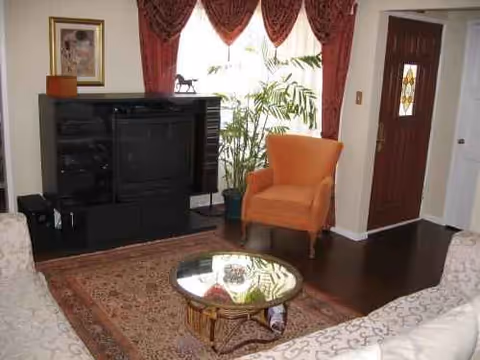 A cozy senior living lounge with orange chair, sofa, rug, plants, and a TV cabinet.