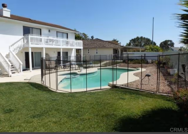 Outdoor view of a residential backyard featuring a swimming pool enclosed by a safety fence. The area includes a two-story white house with a balcony and stairs leading to the ground level. The yard has a well-maintained lawn and some landscaping with plants and gravel along the fence.