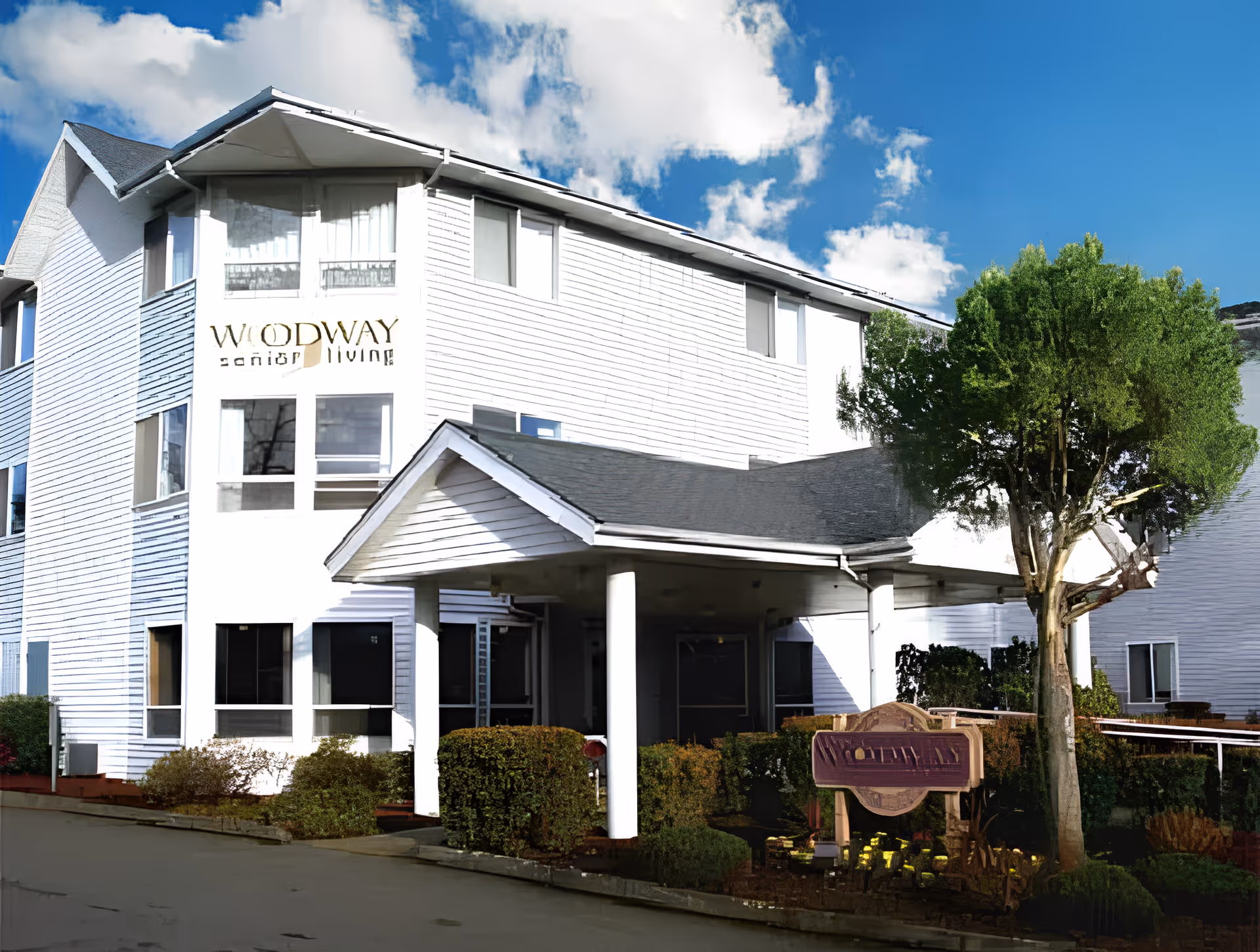 White multi-story senior living building with a covered entrance, landscaping, and a 'Woodway Senior Living' sign.