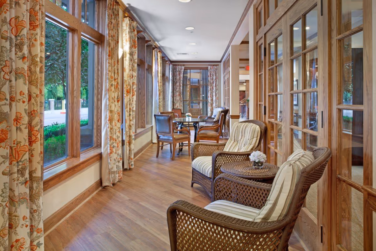 A bright and cozy sitting area with large windows covered by floral curtains, wicker chairs with striped cushions, wooden tables, and additional wooden chairs further down the hallway. The space has wooden flooring and wood-framed glass doors on the right side.