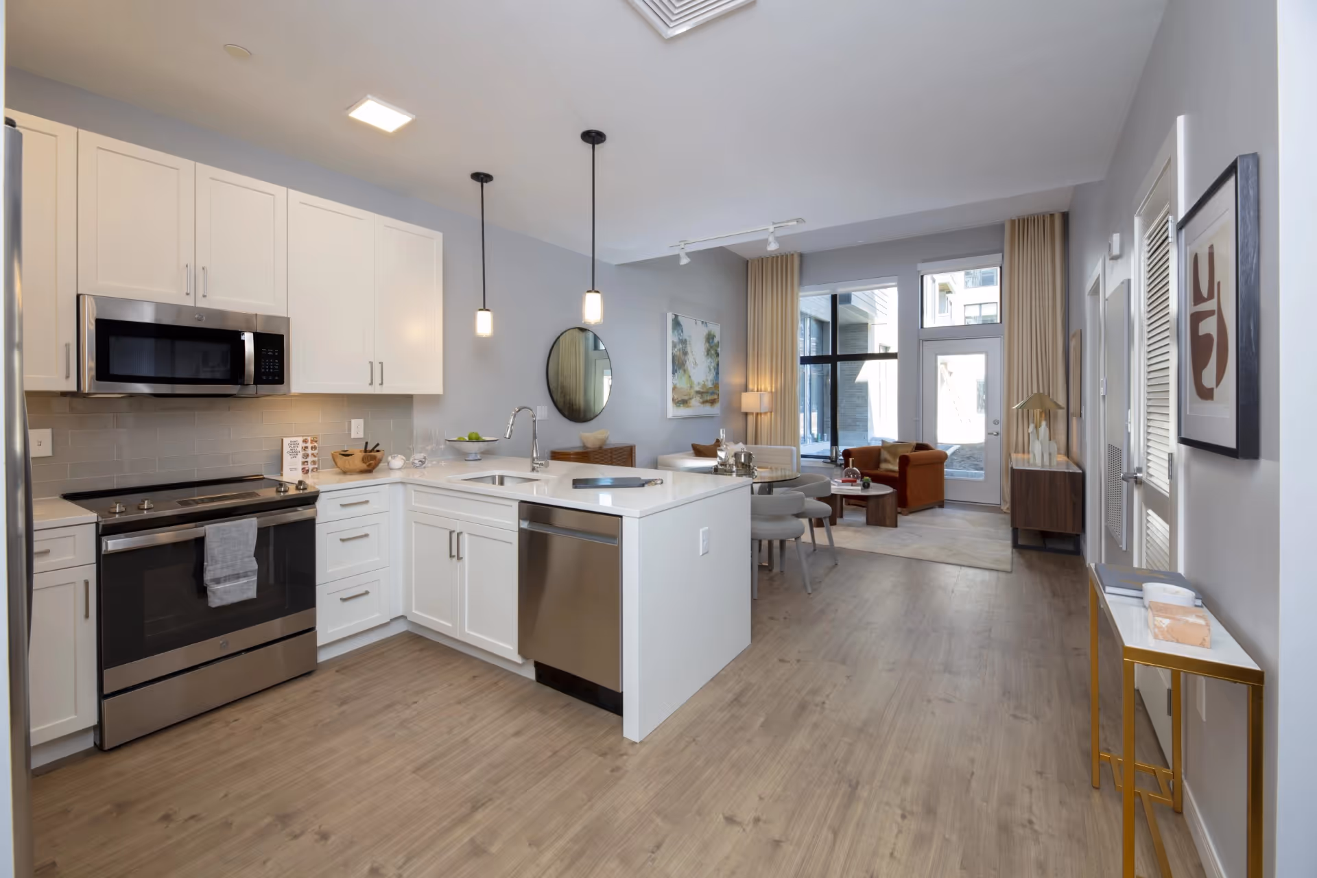 Modern open-concept kitchen and living area with white cabinetry, stainless steel appliances including a stove, microwave, and dishwasher, a kitchen island with a sink and two pendant lights above. The living area has a round dining table with four chairs, a brown armchair, a white sofa, a floor lamp, and large windows with beige curtains letting in natural light.