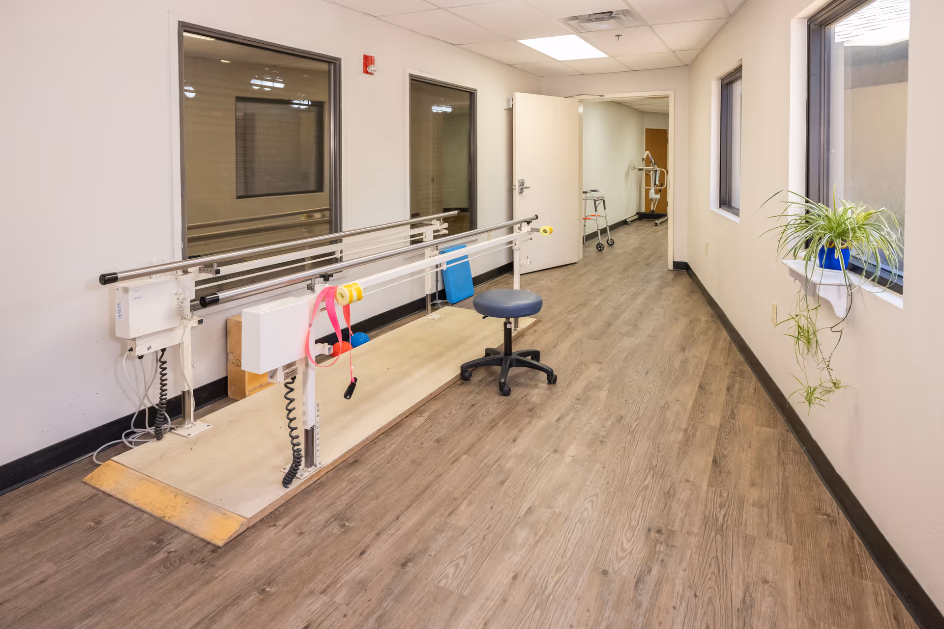 A rehabilitation hallway with parallel bars for physical therapy, a blue stool on wheels, a walker in the background, and a potted plant on a wall-mounted shelf near a window.
