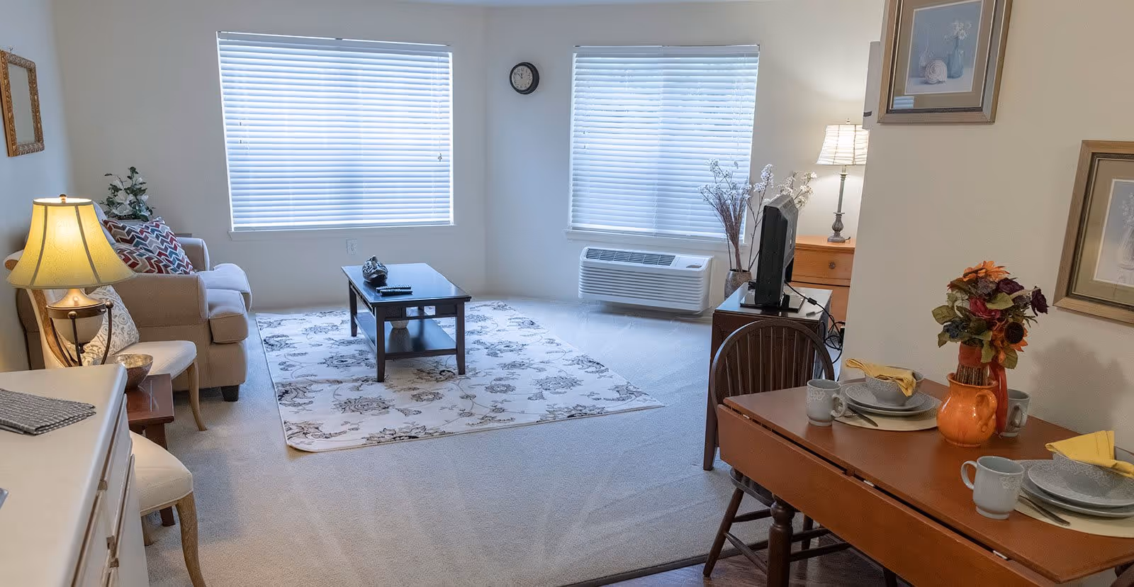 A bright and cozy living room area with two large windows covered by blinds. The room features a beige sofa with patterned cushions, a wooden coffee table on a floral rug, a TV on a wooden stand, and a small dining table set for two with plates, mugs, and a vase of flowers. There are two lamps providing warm lighting and framed artwork on the walls.