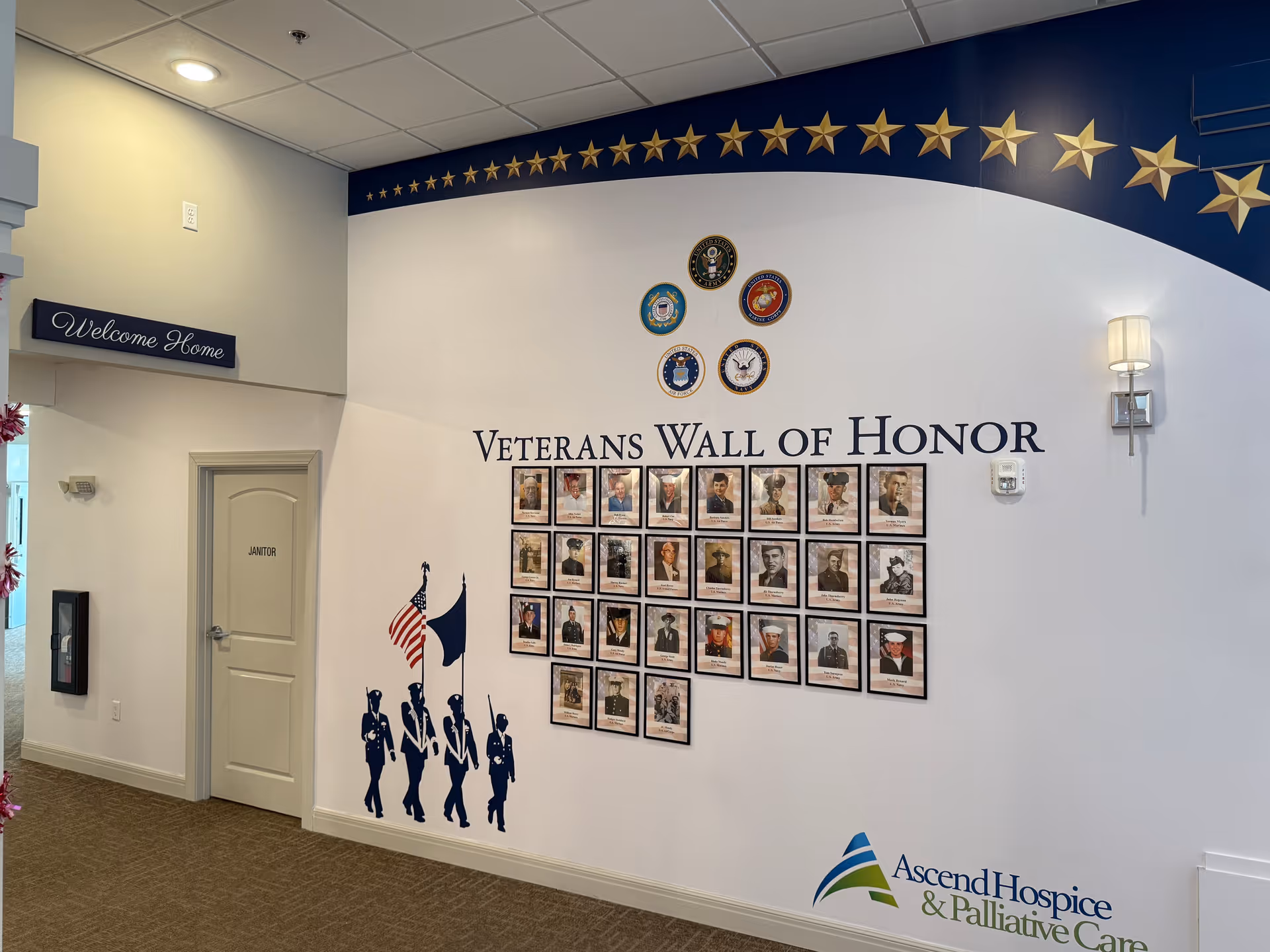 Interior wall display titled 'Veterans Wall of Honor' featuring framed photos of veterans, military service emblems, and a silhouette of a color guard holding flags. The wall is decorated with a blue border and gold stars along the top. To the left, there is a door labeled 'Janitor' and a sign that says 'Welcome Home'. The floor is carpeted and the lighting is bright.