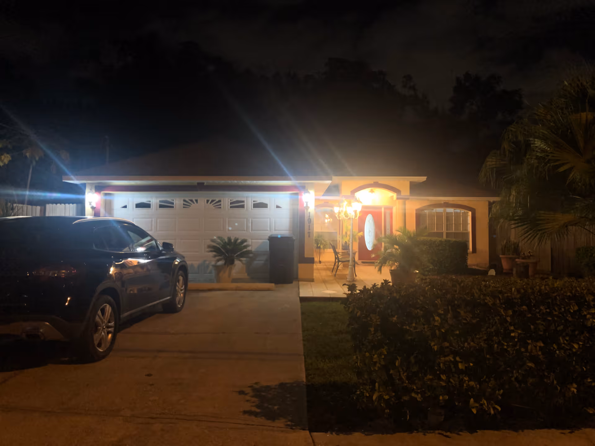 Front exterior of a single-story house at night with a lit porch, closed garage, and a car parked in the driveway.