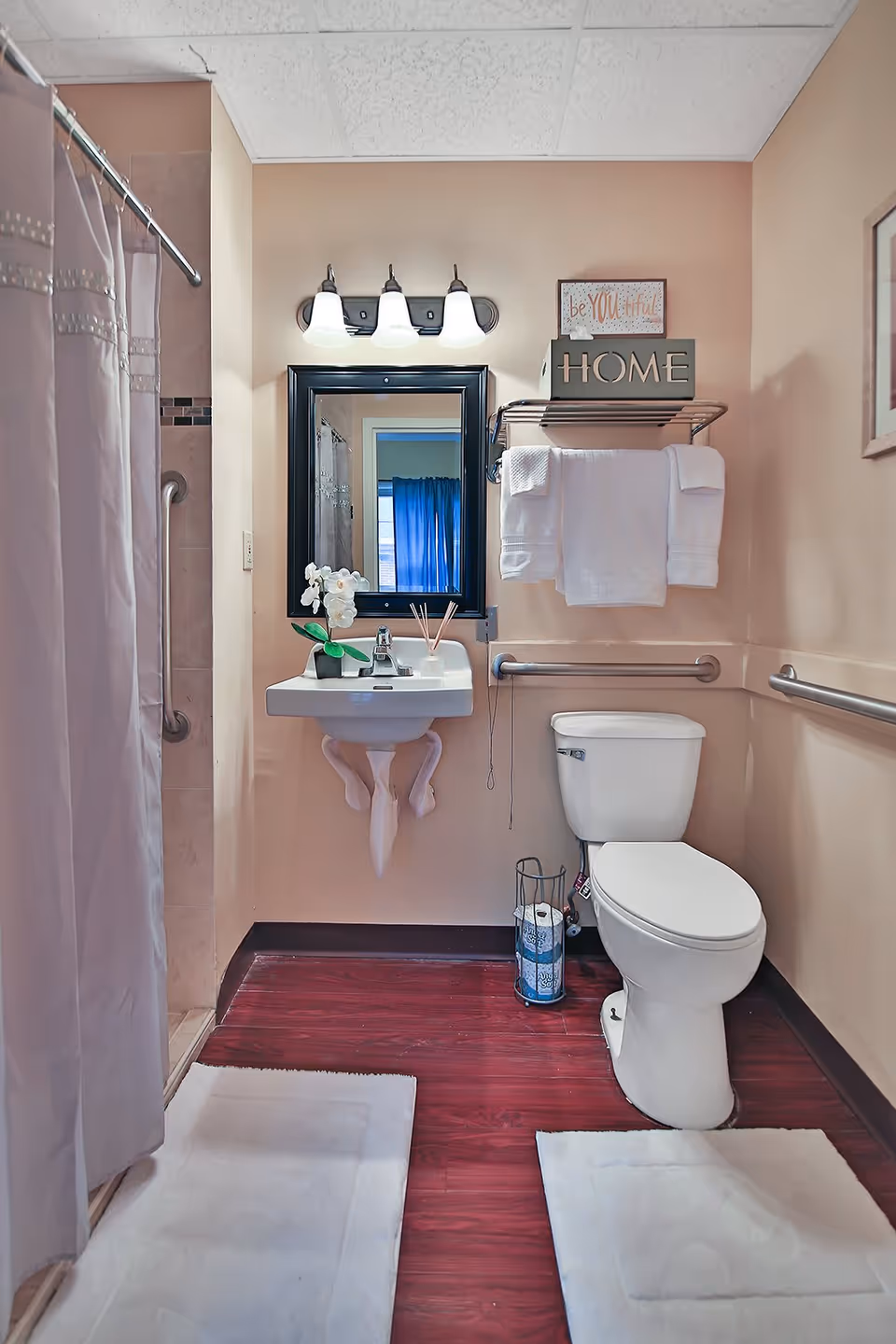 A clean and accessible bathroom with a white toilet, a wall-mounted sink with decorative legs, a mirror above the sink with three light fixtures, and a shower with a curtain on the left. There are grab bars on the walls near the toilet and shower. White towels are neatly folded on a metal rack above the toilet, and a small decorative sign that says 'HOME' and another sign that says 'be YOU tiful' are placed on the rack. The floor has dark wood-like flooring with two white bath mats.