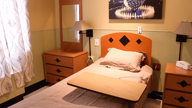 A single bed in a room with a wooden headboard featuring diamond-shaped decorations. The bed has a beige pillow and blanket, and a wooden overbed table is positioned on top. On either side of the bed are wooden nightstands with matching diamond-shaped decorations, one with a white telephone. A wall-mounted lamp is above each nightstand. A dresser with a mirror is next to the bed, and a window with patterned curtains lets in natural light. A framed picture of a globe and candles hangs on the wall above the bed.