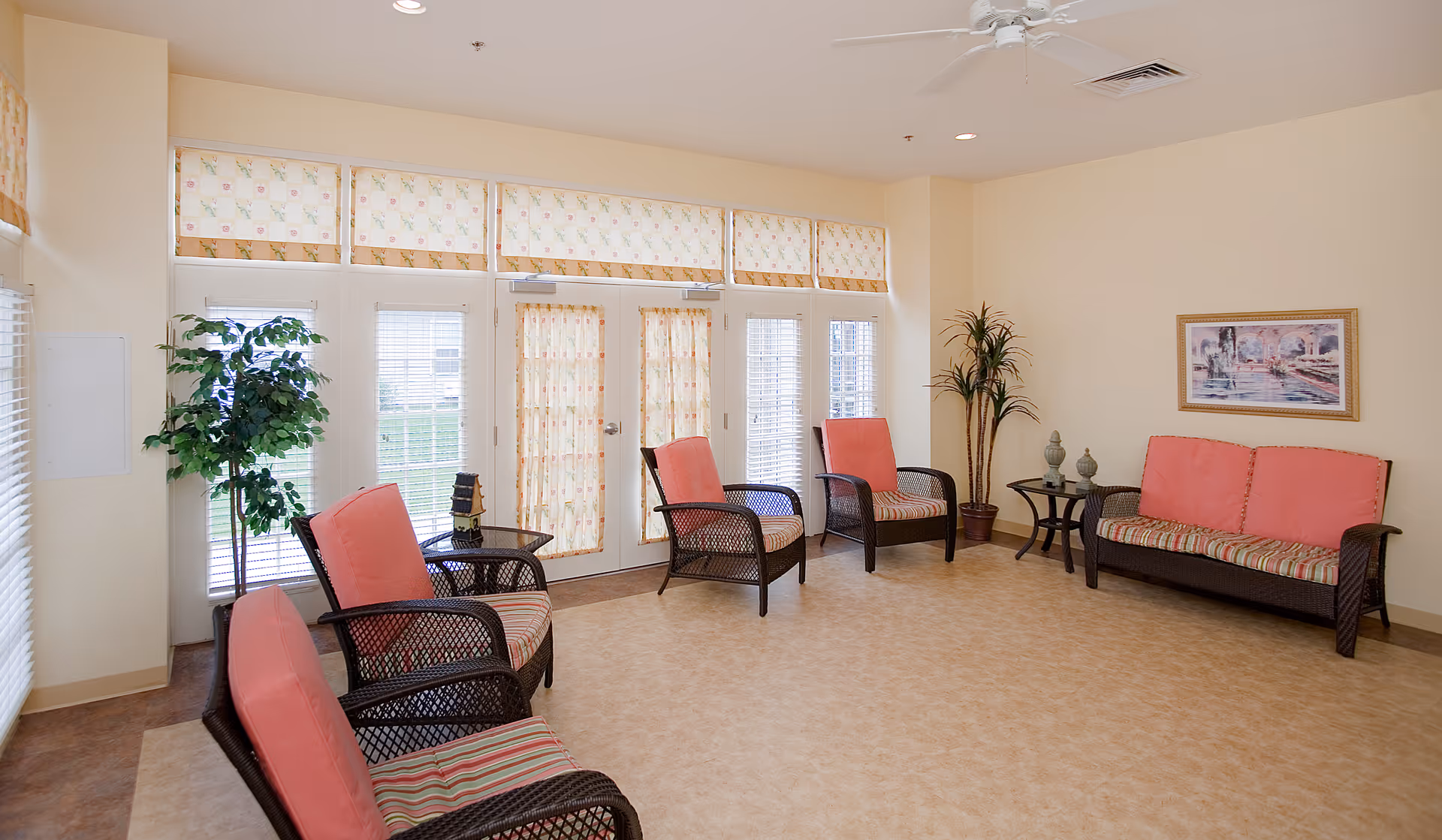 A bright and spacious living room area with beige walls and tiled floor. The room features several wicker chairs and a sofa with coral cushions and striped seat covers arranged around small glass-top tables. Large windows and glass doors with floral curtains allow natural light to enter. There are two potted plants and a framed painting on the wall.