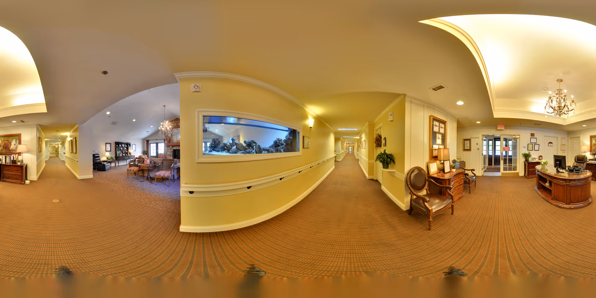 Panoramic view of a senior living facility interior hallway with beige walls and carpet. On the left side, there is a cozy sitting area with chairs, tables, and a fireplace. In the center, a long hallway with handrails and a large built-in aquarium is visible. On the right side, there is a reception desk with chairs, lamps, and framed pictures on the walls. The ceiling features recessed lighting and a chandelier near the entrance doors.