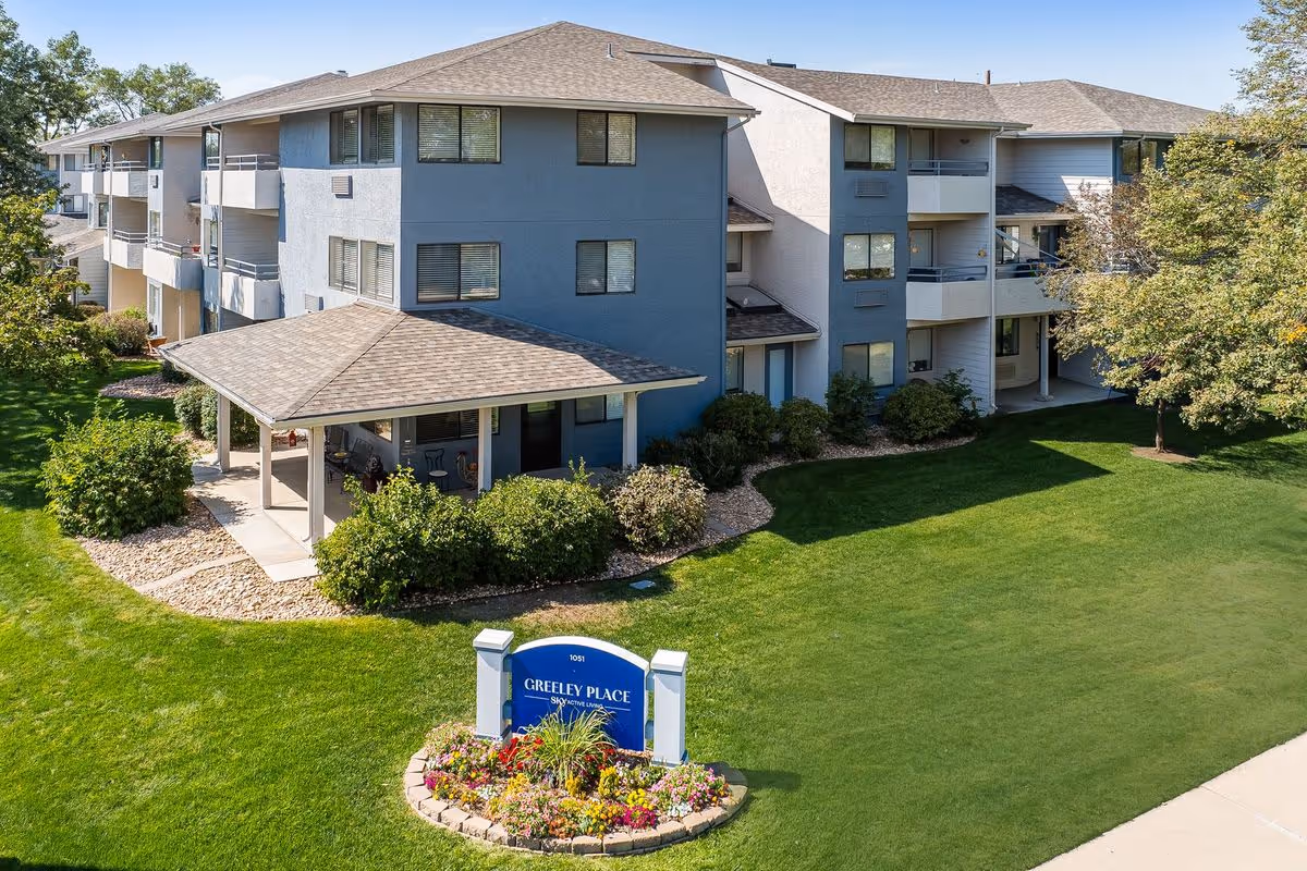 Exterior view of a multi-story senior living facility building with balconies, surrounded by well-maintained green lawns, bushes, and trees. A covered patio area is visible on the left side of the building. In the foreground, there is a blue sign with white text that reads 'Greeley Place | Sky Active Living' surrounded by a flower bed.