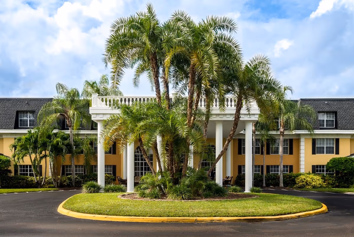 Front exterior view of Volante Senior Living of Delray Beach showing a yellow two-story building with white columns at the entrance, surrounded by palm trees and greenery under a partly cloudy sky.