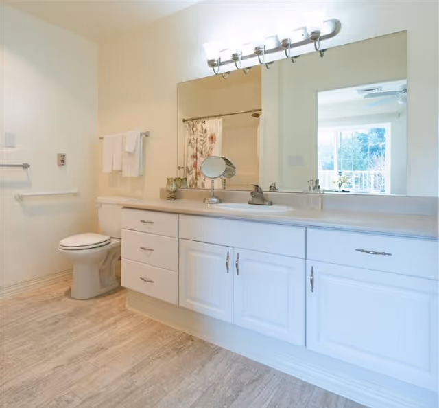 Bright bathroom with a large mirror above a white vanity with multiple drawers and cabinets. A single sink with a faucet is centered on the countertop. To the left, there is a toilet and a towel rack with white towels. The shower curtain with a floral pattern is visible in the mirror reflection. The floor has light-colored tiles and the walls are painted a soft beige.