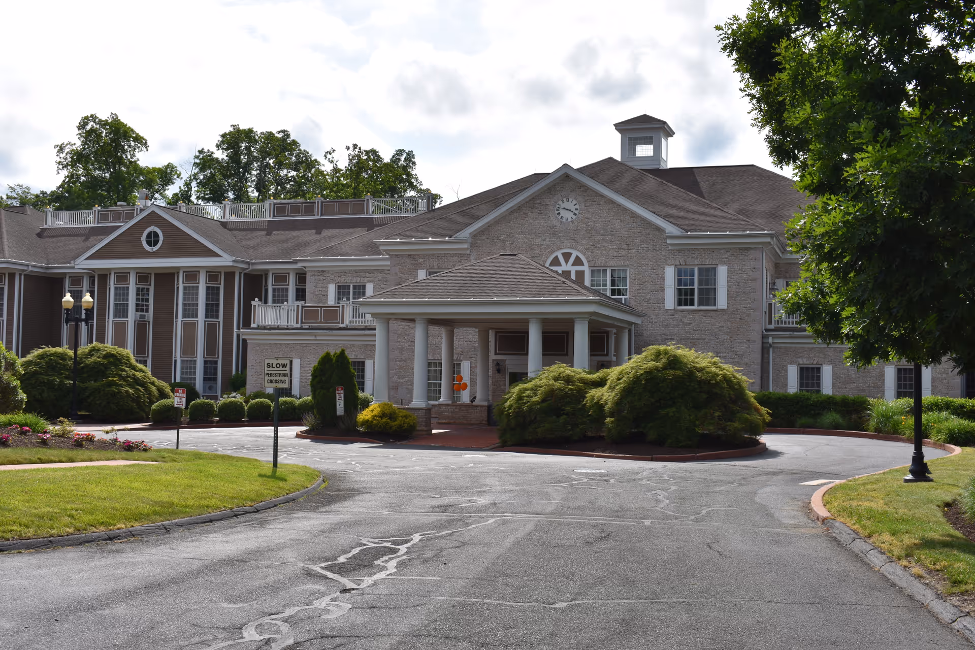 Front exterior view of a large senior living facility building with a circular driveway, manicured bushes, and trees. The building has a brick facade with multiple windows, a clock above the entrance, and a covered portico supported by columns.