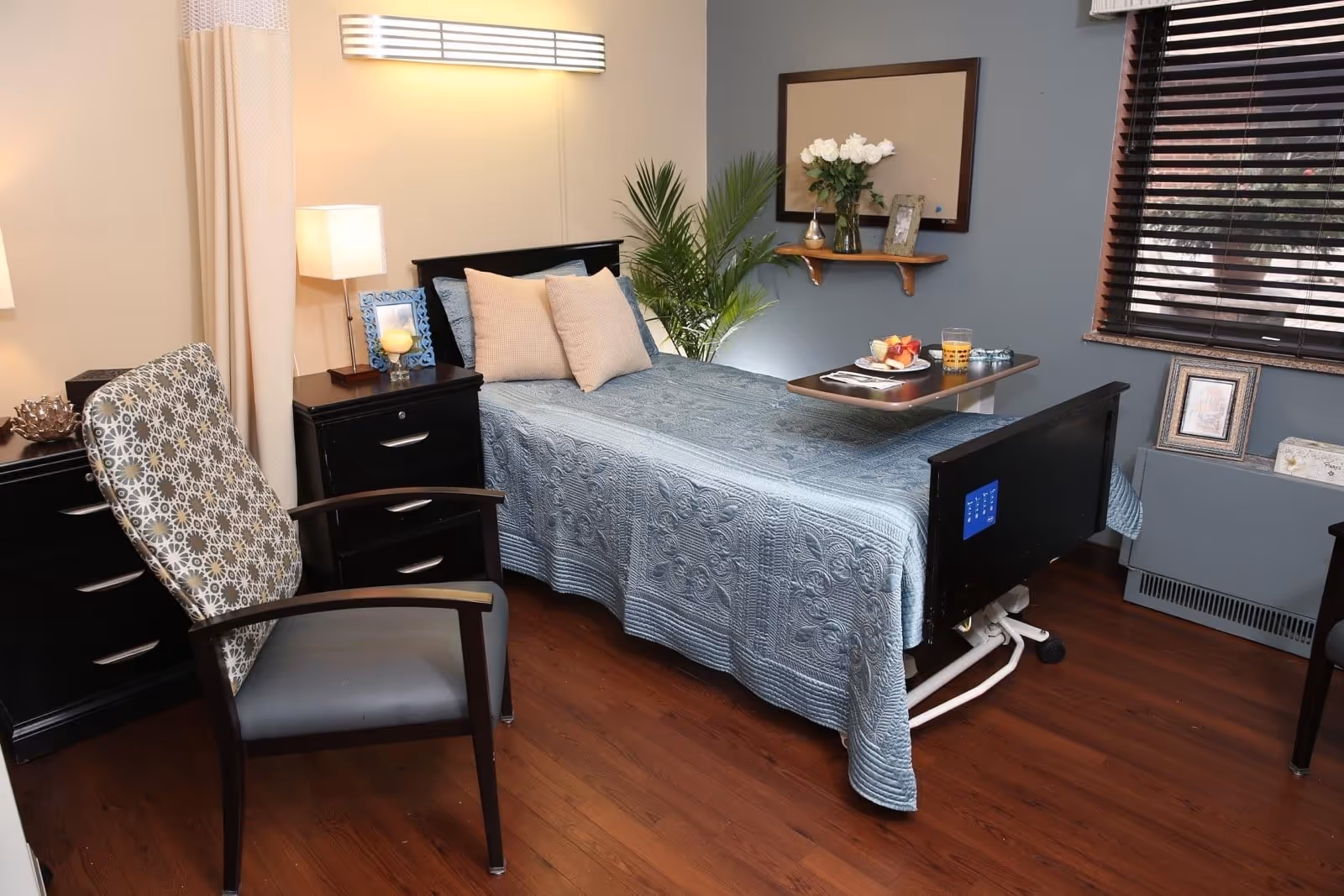 A cozy senior resident bedroom with a single adjustable bed, overbed table, armchair, and bedside drawers.