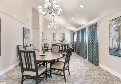 A bright dining room with round tables and cushioned chairs arranged neatly. The room features patterned carpet flooring, large windows with blue patterned curtains, and modern chandeliers hanging from a vaulted ceiling. There are framed paintings on the walls adding to the decor.