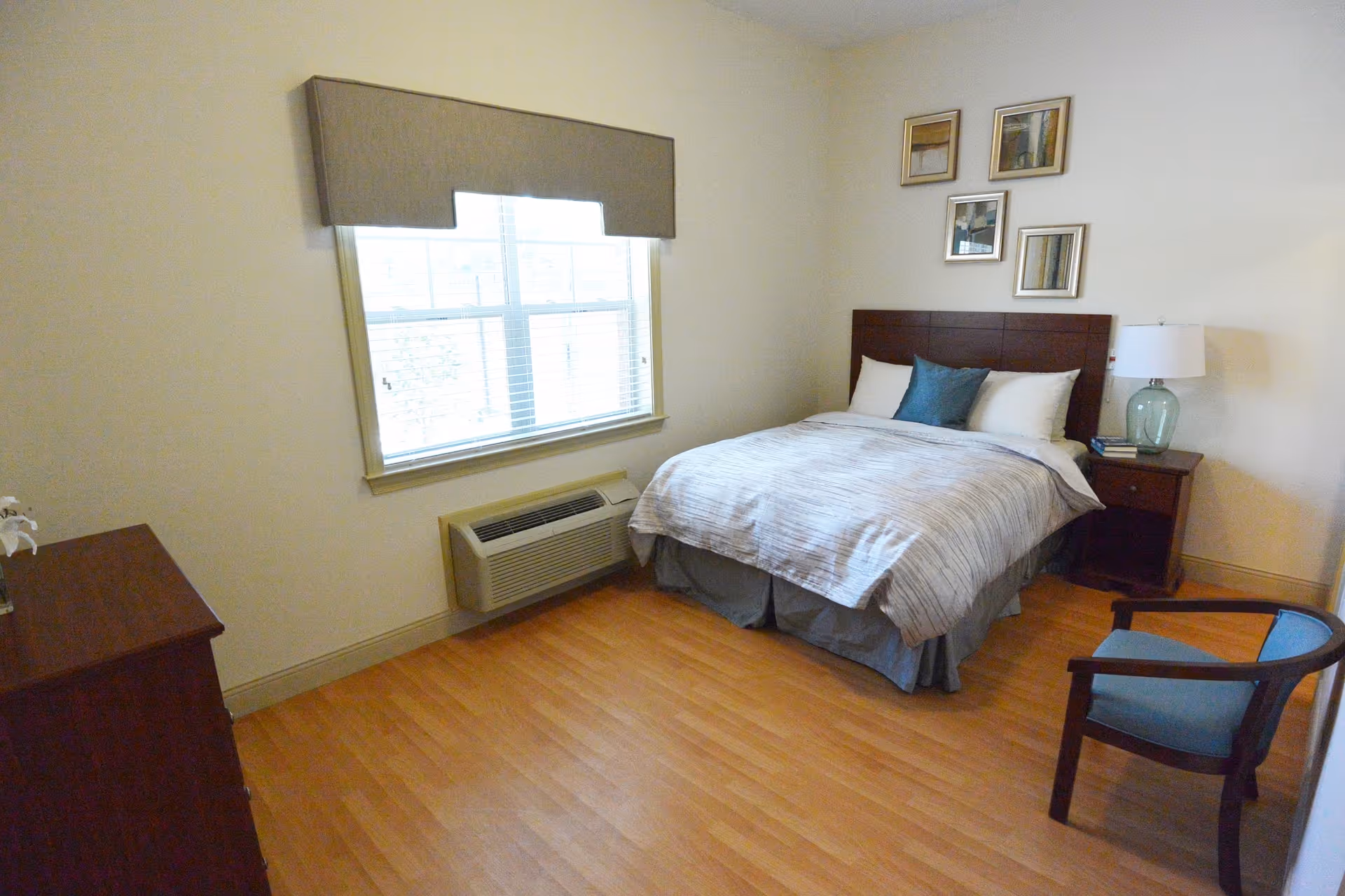A simple bedroom with a bed featuring white and gray bedding and a blue accent pillow. There is a wooden headboard, a nightstand with a lamp and books, a wooden dresser, a blue cushioned chair, and a window with blinds and a valance. The floor is light wood, and there are four framed pictures on the wall above the bed.