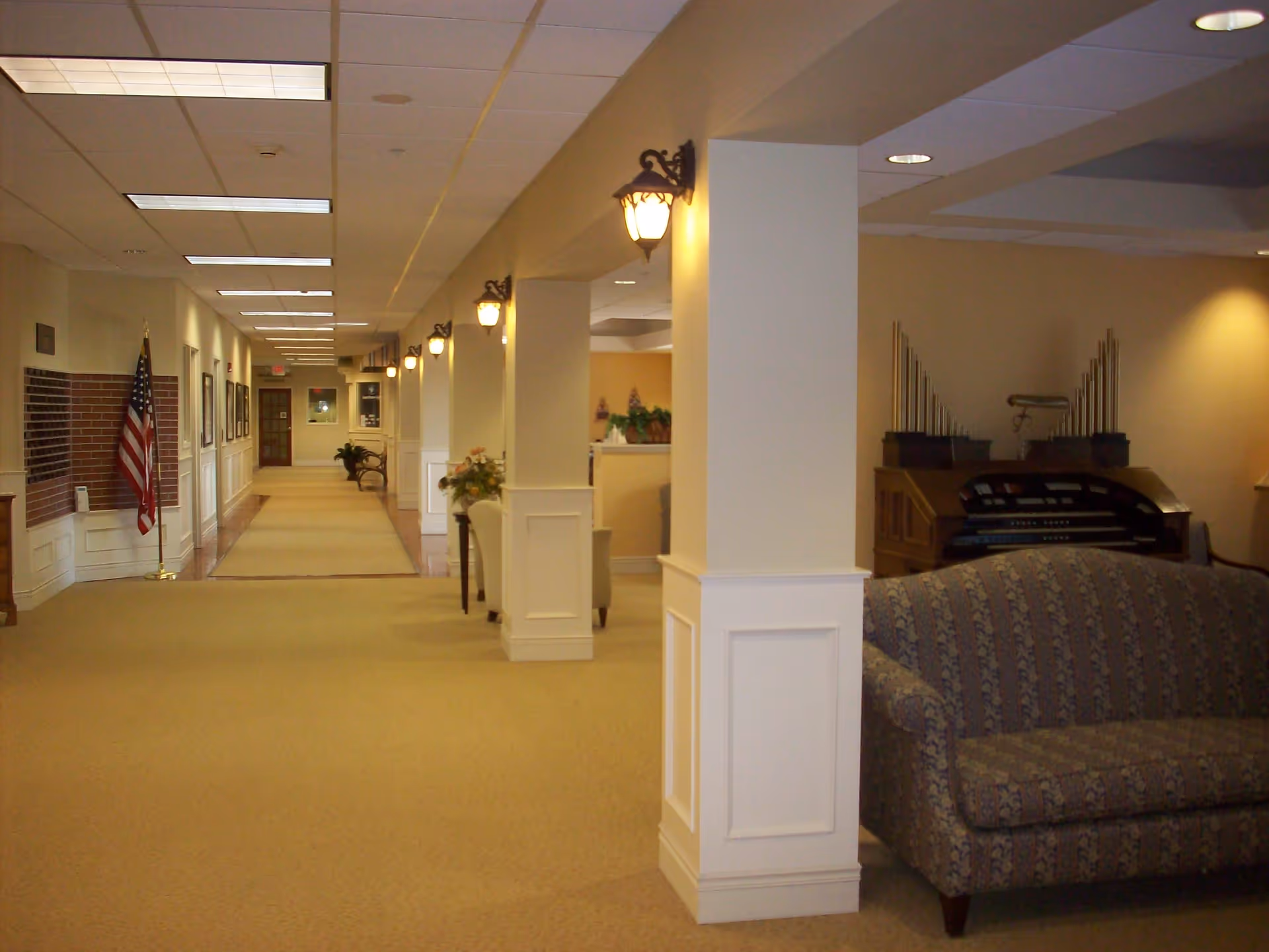 Interior hallway of a senior living facility with beige carpet and walls, white pillars with wall-mounted lamps, an American flag on the left, framed pictures on the wall, a patterned couch on the right, and a large organ in the background.