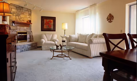 Cozy living room featuring a stone fireplace, a white striped sofa, an armchair, and a glass coffee table, with natural light coming from a window.