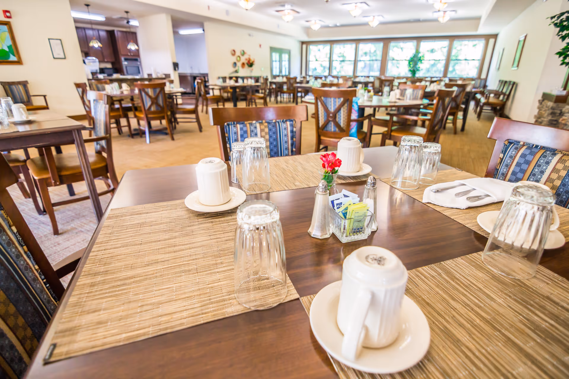 A bright and spacious dining room in a senior living facility with multiple wooden tables and chairs. Tables are set with placemats, upside-down glasses, white coffee mugs on saucers, silverware wrapped in napkins, and a small centerpiece with a red flower and condiments. Large windows in the background let in natural light, and the room has a warm, inviting atmosphere.