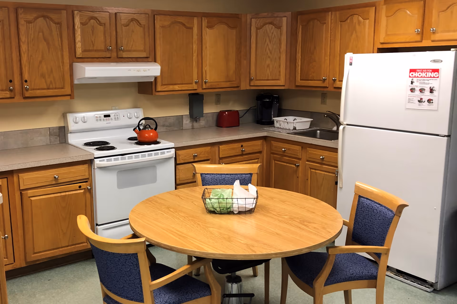 A kitchen area with wooden cabinets, a white electric stove with an orange kettle on top, a white refrigerator with a first aid for choking poster, a countertop with a red toaster and a coffee maker, and a round wooden table with four chairs that have blue cushioned seats and backs. A wire basket with some items is on the table.