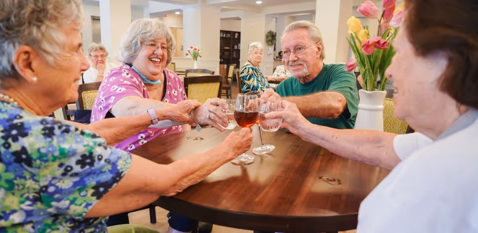 Four elderly people sitting around a wooden table in a well-lit room, smiling and clinking their glasses together in a toast. The room has light-colored walls, decorative columns, and a vase with flowers on the table. Other elderly individuals are visible in the background seated at tables.