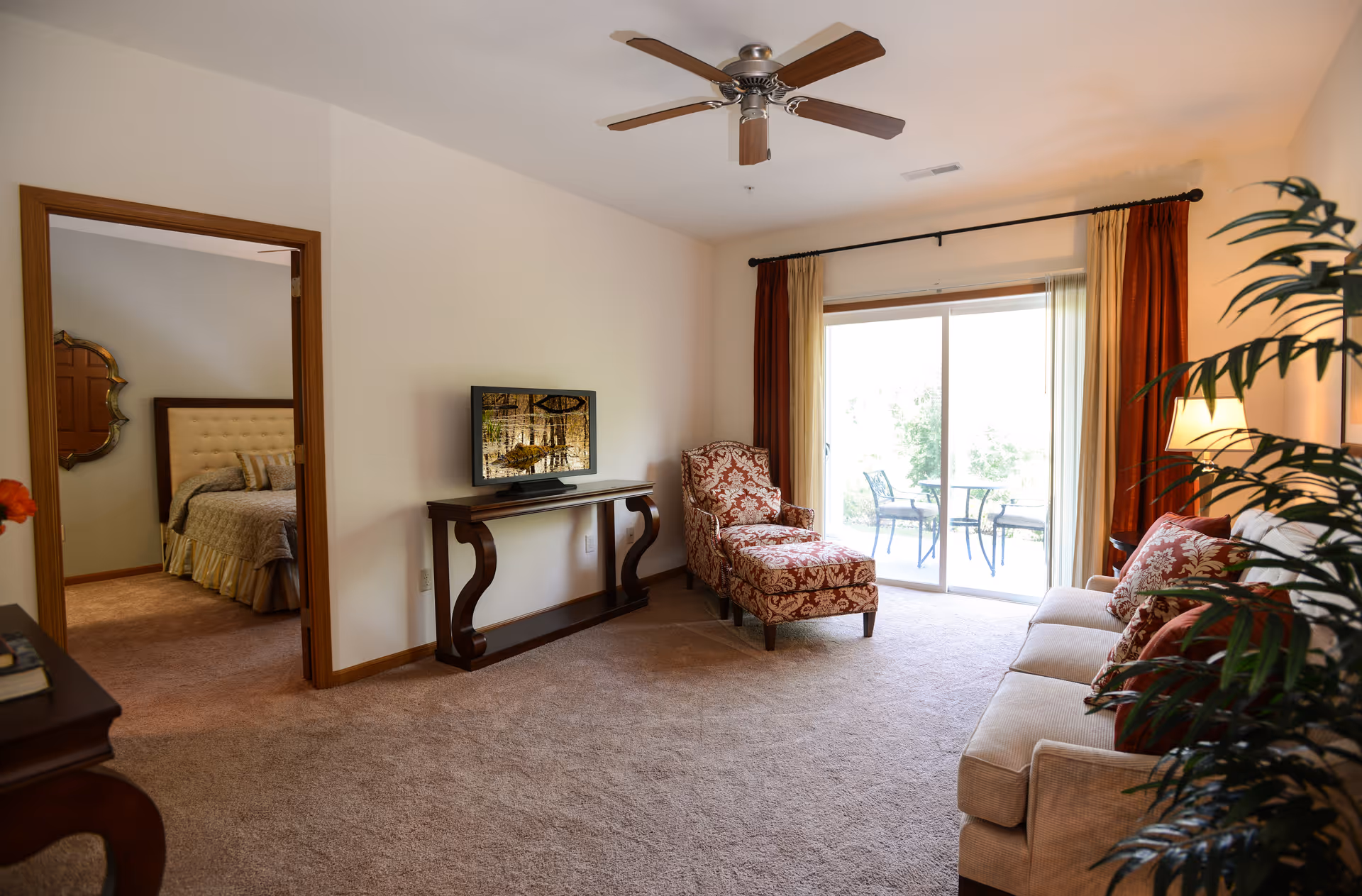 A cozy living room with a beige sofa adorned with patterned cushions, a matching armchair with an ottoman, a flat-screen TV on a wooden console table, a ceiling fan, and sliding glass doors leading to an outdoor patio with a table and chairs. A doorway on the left reveals a bedroom with a bed and decorative mirror.